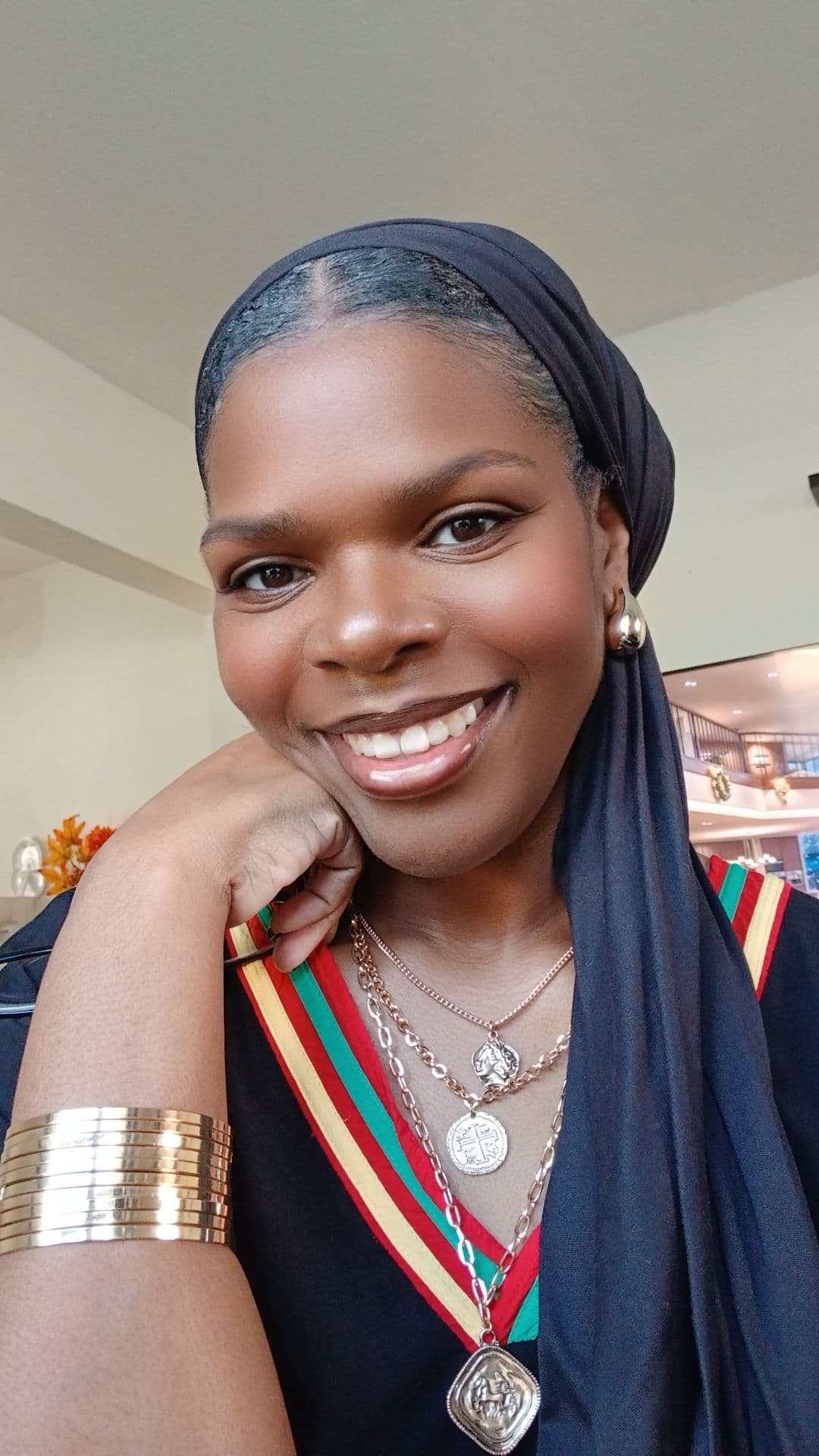 Smiling Black woman wearing a black headwrap, layered gold necklaces, and a striped top.
